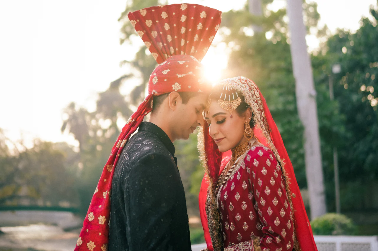 Bride and groom in red wedding attire touch foreheads at golden hour, the sun flaring between them