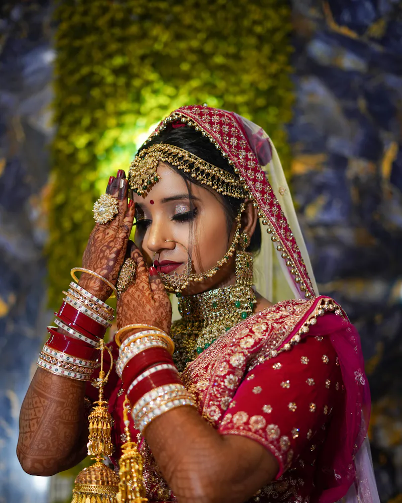 Bride in red and gold couture, mehndi-covered hand raised to her face